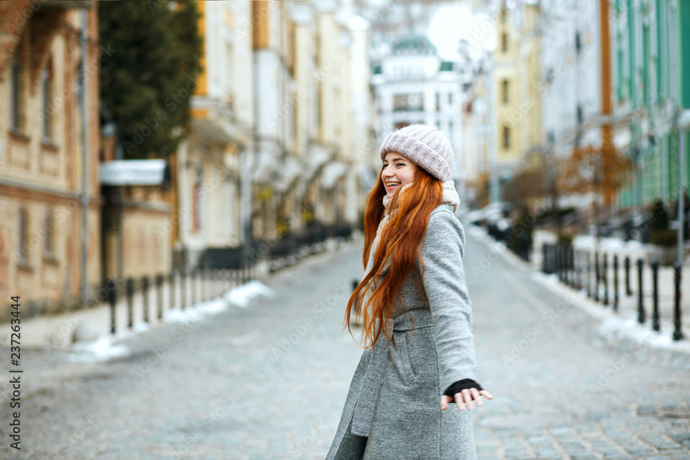 Pretty red haired model wearing stylish winter outfit walking in city, spinning around. Empty space