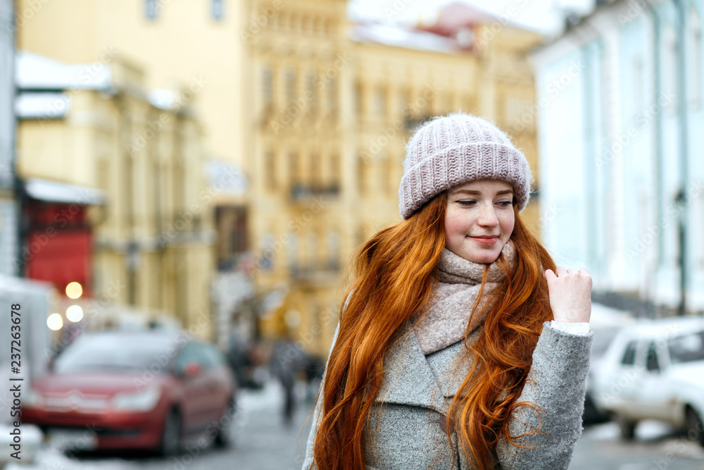Street portrait of lovely redhead girl with long hair wearing warm winter apparel posing at the street. Empty space