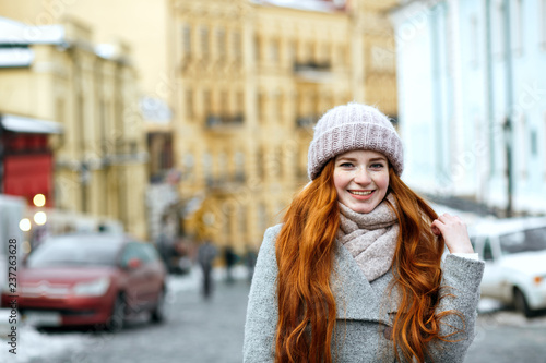 Street portrait of positive redhead model with long hair wearing warm winter apparel posing at the street. Empty space
