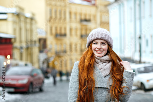 Street portrait of cheerful redhead model with long hair wearing warm winter apparel posing at the street. Empty space