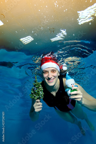 A girl swimming coach is swimming underwater with a snowman toy and a little Christmas tree in her hand in Santa's red cap, looking at me and smiling on a blue background. Portrait