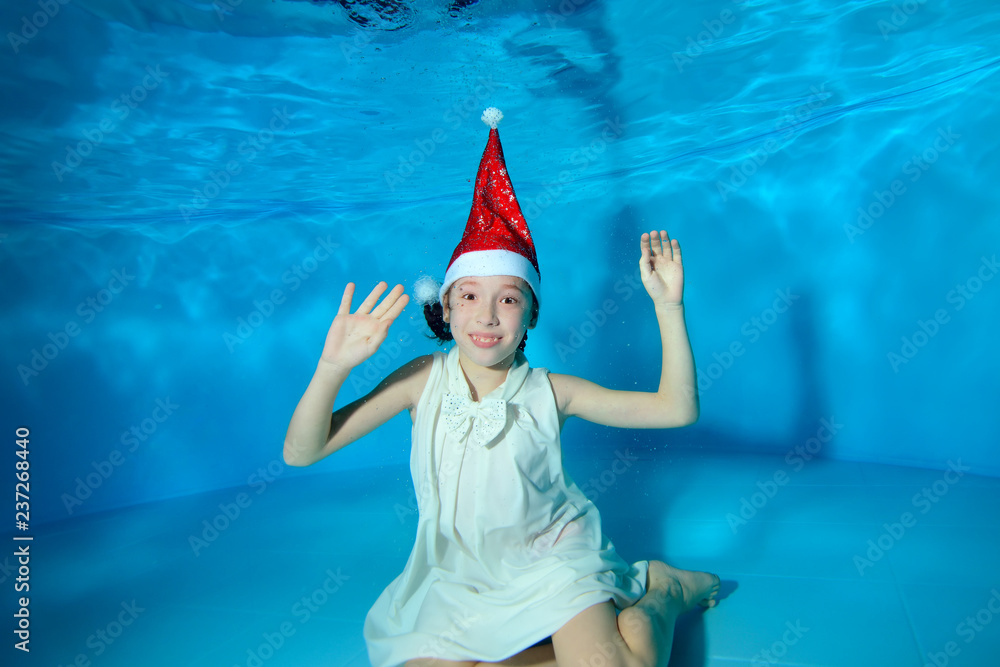 Obraz premium A little girl is sitting underwater at the bottom of the pool in a red Santa hat and a white dress on a blue background. She raised her hands up, looks at the camera and smiles. Portrait