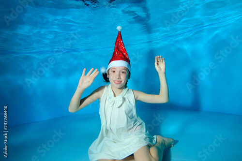 A little girl is sitting underwater at the bottom of the pool in a red Santa hat and a white dress on a blue background. She raised her hands up, looks at the camera and smiles. Portrait