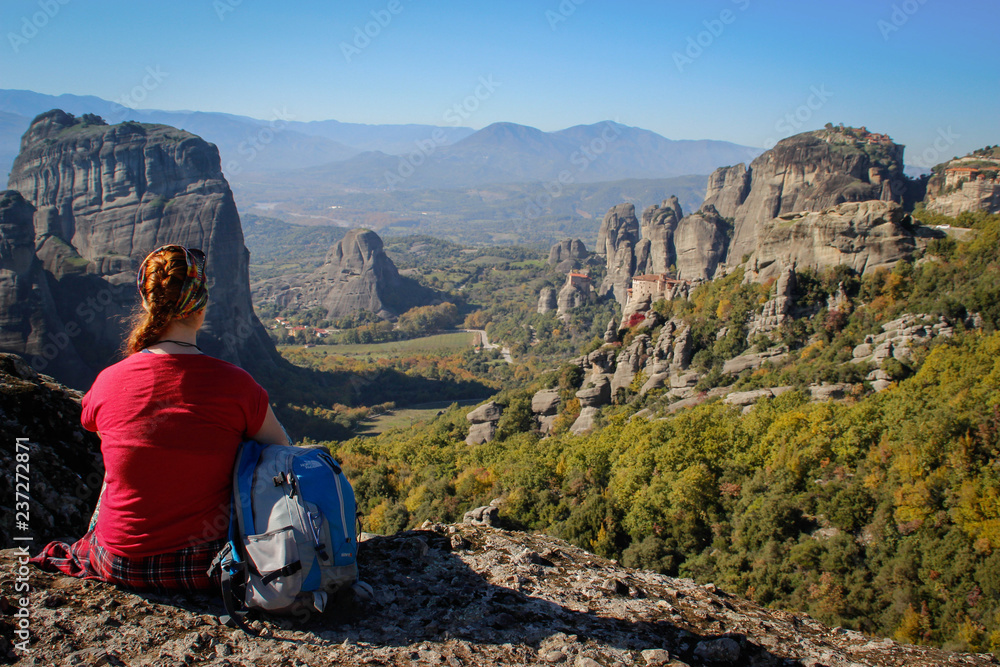 Naklejka premium A traveler girl in a red T-shirt and a blue backpack sits high against the rocks of the Meteora temple complex in Greece on a bright summer sunny day.