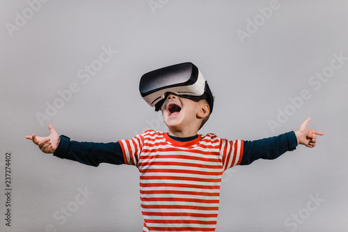Excited kid with hands spread having fun with VR glasses. Portrait of happy child wearing virtual reality headset against grey background.