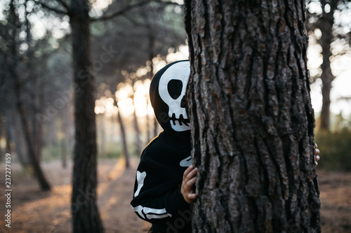 Kid in skeleton costume behind the tree in forest. photo