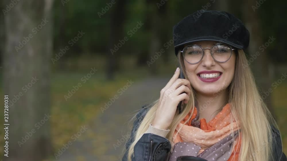 Young adorable blonde woman using smartphone for call at city park cold season, holding coffee cup, smiling into the camera. Close up, shallow depth of field. Young adorable blonde woman using smartphone for call at city park cold season, holding coffee cup, smiling into the camera. Close up, shallow depth of field.
