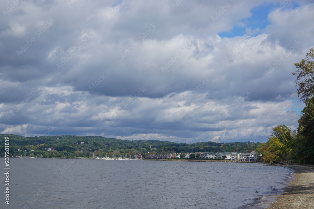 Croton-On-Hudson, New York, USA: View of houses and a marina on the Hudson River under a cloud-filled sky, from a pebble beach  in Croton Point Park.