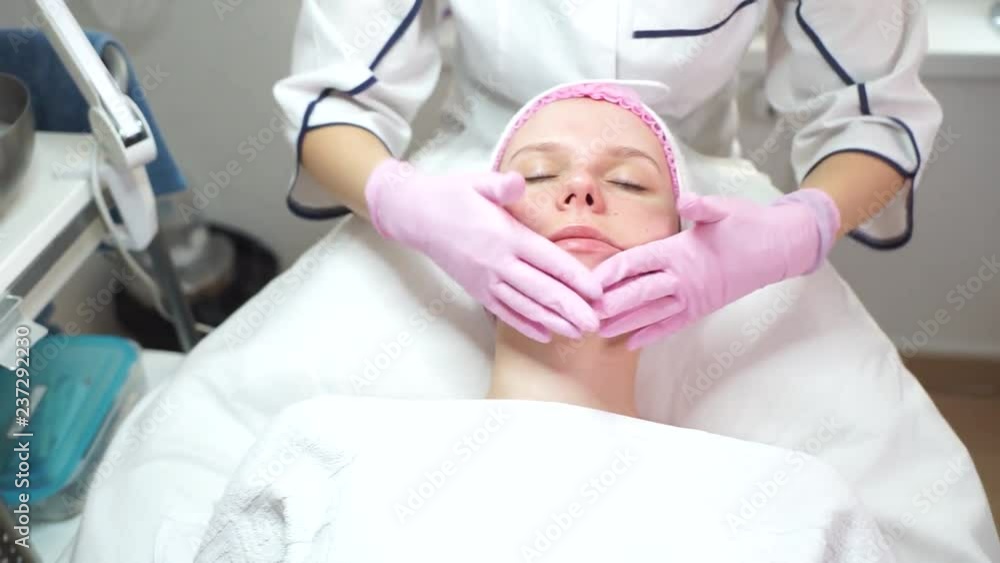 Moisturizing skin for a European girl. Beautiful girl in a beauty salon. Cosmetologist makes anti-aging massage. The beautician applies the product to the woman's face. Moisturizing skin for a European girl. Beautiful girl in a beauty salon. Cosmetologist makes anti-aging massage. The beautician applies the product to the woman's face.