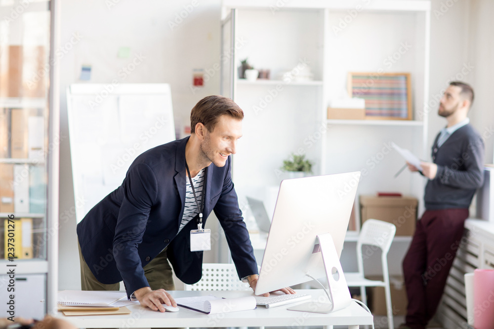 Side view portrait of smiling businessman using computer standing at desk in office, copy space Side view portrait of smiling businessman using computer standing at desk in office, copy space