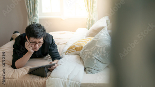 Young Asian business man using digital tablet and pen while lying on bed in cozy bedroom. Home living lifestyle with modern electronic gadget concept