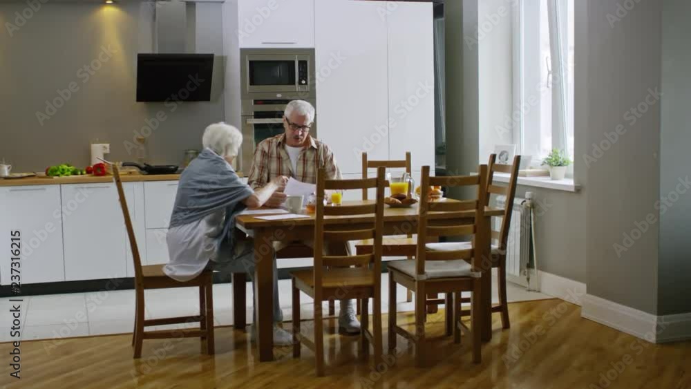 Wide shot of old couple sitting at kitchen table, looking through some papers and discussing them Wide shot of old couple sitting at kitchen table, looking through some papers and discussing them