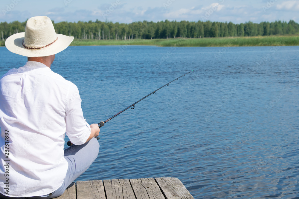 man, on the background of nature, sits on the pier with a fishing rod in his hands, view from the back man, on the background of nature, sits on the pier with a fishing rod in his hands, view from the back