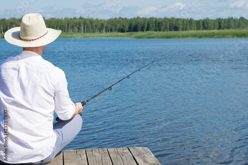 man, on the background of nature, sits on the pier with a fishing rod in his hands, view from the back