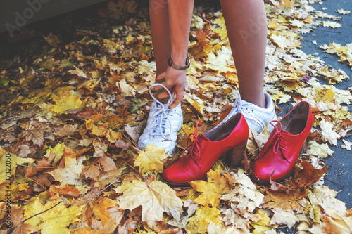 Young woman putting on white sports shoes for active walking in park. Legs of single girl in run snickers on the road among falling yellow maple leaves. Red woman boots wiht high heels. Autumn day.