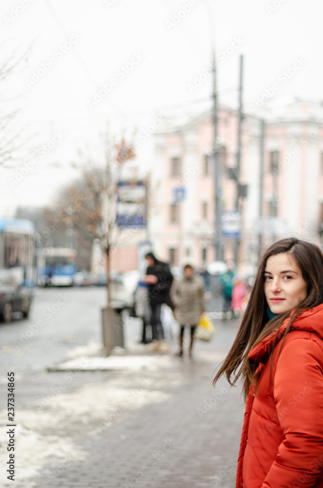 beautiful brunette in a warm red coat or down jacket on a cold, gloomy street poses with a smile for a photo, an attractive woman in a stylish woolen coat in the winter on the street with a smile