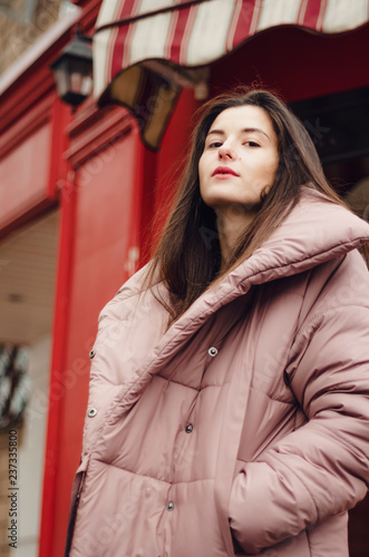 beautiful brunette in a warm pink coat on a cold, gloomy street poses with a smile for a photo, an attractive woman in a stylish woolen coat in the winter on the red background on street