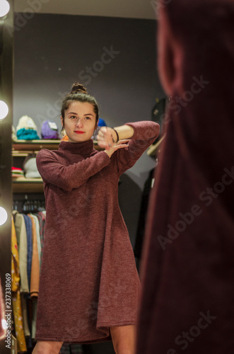 Woman trying clothing looking in mirror adjusting dress. Beautiful young brunette girl. Shopping, fashion, style, wear and people concept - happy woman in front of the mirror at clothing store