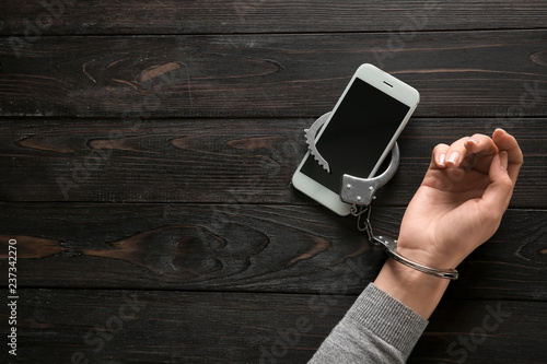 Female hand in handcuffs with mobile phone on wooden background. Concept of addiction