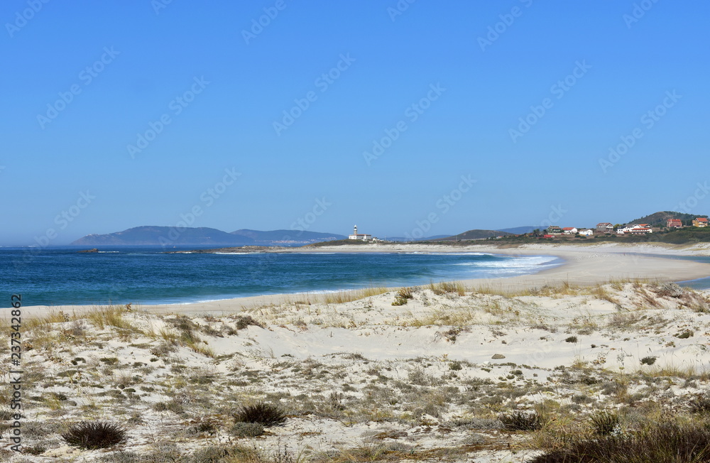 Beach with lake, vegetation in sand dunes and lighthouse. Clear water with waves and foam. Green, blue and turquoise colours. Blue sky, sunny day. Galicia, Spain.