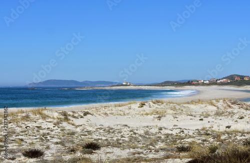 Beach with lake  vegetation in sand dunes and lighthouse. Clear water with waves and foam. Green  blue and turquoise colours. Blue sky  sunny day. Galicia  Spain.