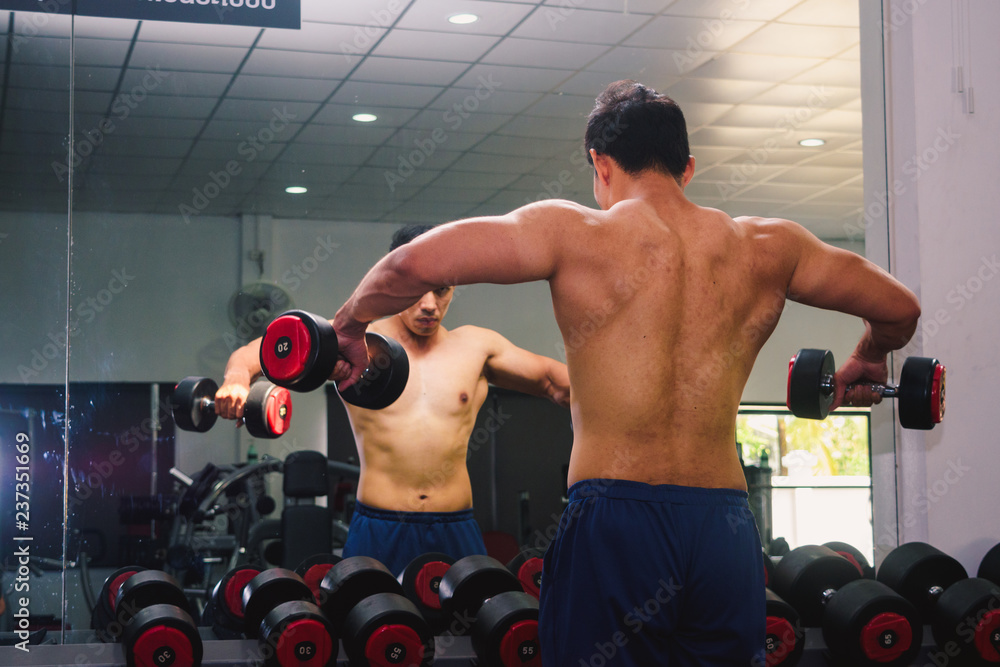 An Asian man lifts a dumbbell in front of a mirror in a gym. Exercise in gym concept An Asian man lifts a dumbbell in front of a mirror in a gym. Exercise in gym concept