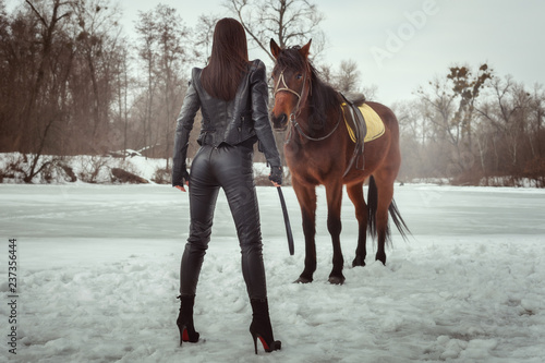 Woman in a black leather suit is standing with her back. Before her stands a horse