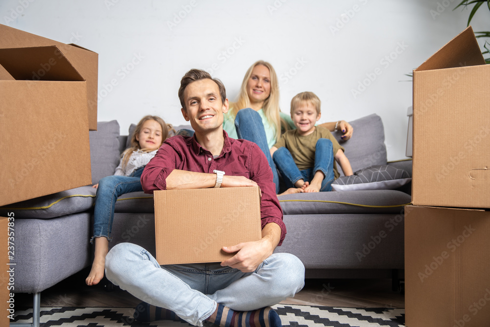 Photo of man sitting on floor and women with boy and girl sitting on gray sofa among cardboard boxes Photo of man sitting on floor and women with boy and girl sitting on gray sofa among cardboard boxes