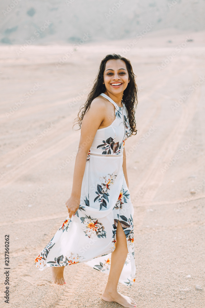 Young indian woman in white long dress walking in the desert with the mountains on background Young indian woman in white long dress walking in the desert with the mountains on background
