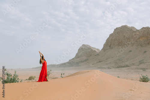 Young beautiful indian woman in red dress standing in the desert with her hands above the head photo