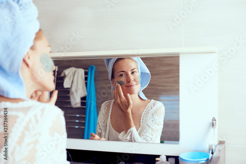 Young pretty woman in bathrobe and with towel on head removing facial mask in front of mirror in bathroom. Skin care and beauty concept
