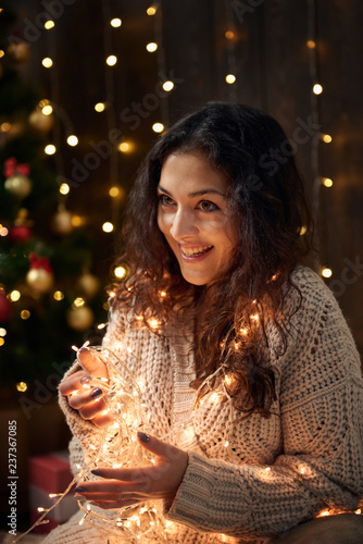 young girl is in christmas lights and decoration, dressed in white, fir tree on dark wooden background, winter holiday concept © soleg