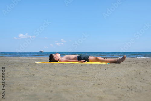 Teen  boy lies on yellow towel and sunbathes on the beach on the sea and sky background. Concept
