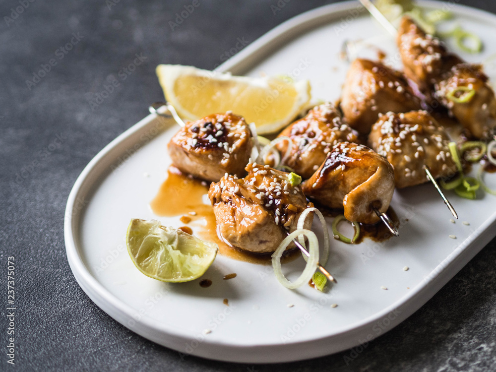 Grilled Asian teriyaki chicken skewers with sesame, leek and citrus juice on a white oval plate on dark grey background Grilled Asian teriyaki chicken skewers with sesame, leek and citrus juice on a white oval plate on dark grey background