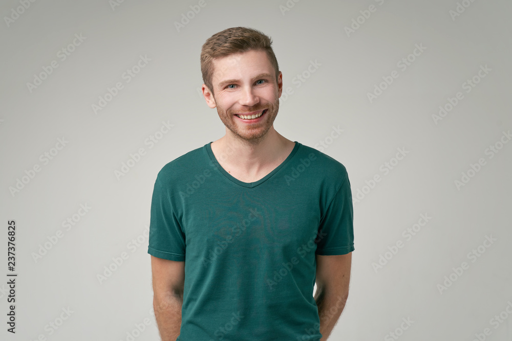 Portrait of enthusiastic man, smiles broadly, laugh joyfully at funny rumor, wears casual green t shirt. Content student has break after classes indoor. Isolated on white background