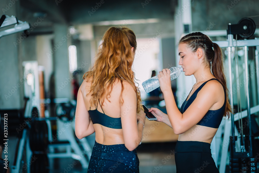 Fit beautiful young woman athletes in fitness clothes relaxing after workout in gym. Couple women standing holding water in bottle and using fitness app smartphone. Healthy, Sport, Lifestyle concept. Fit beautiful young woman athletes in fitness clothes relaxing after workout in gym. Couple women standing holding water in bottle and using fitness app smartphone. Healthy, Sport, Lifestyle concept.