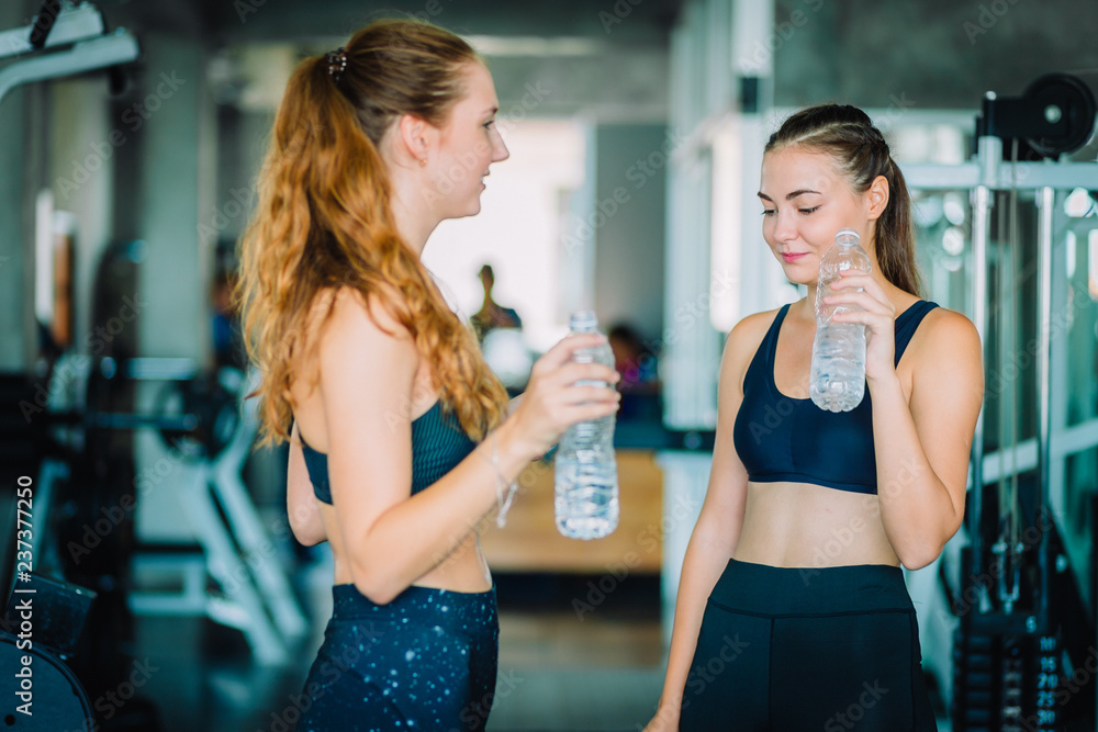 Fit beautiful young woman athletes in fitness clothes relaxing after workout in gym. Couple women standing holding water in bottle. Healthy, Sport, Lifestyle concept. Fit beautiful young woman athletes in fitness clothes relaxing after workout in gym. Couple women standing holding water in bottle. Healthy, Sport, Lifestyle concept.