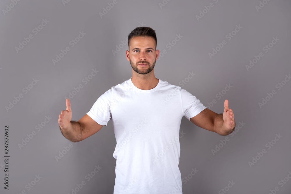 A handsome young man in a white tshirt showing something with his palms in front of a grey background in the studio. A handsome young man in a white tshirt showing something with his palms in front of a grey background in the studio.