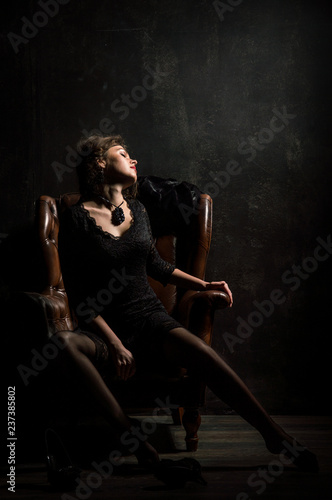 Fototapeta Naklejka Na Ścianę i Meble -  Roaring Twenties. Woman portrait in the style of Gatsby. Low key. A beautiful young woman in a black lace dress is sensually sitting in a brown leather chair.