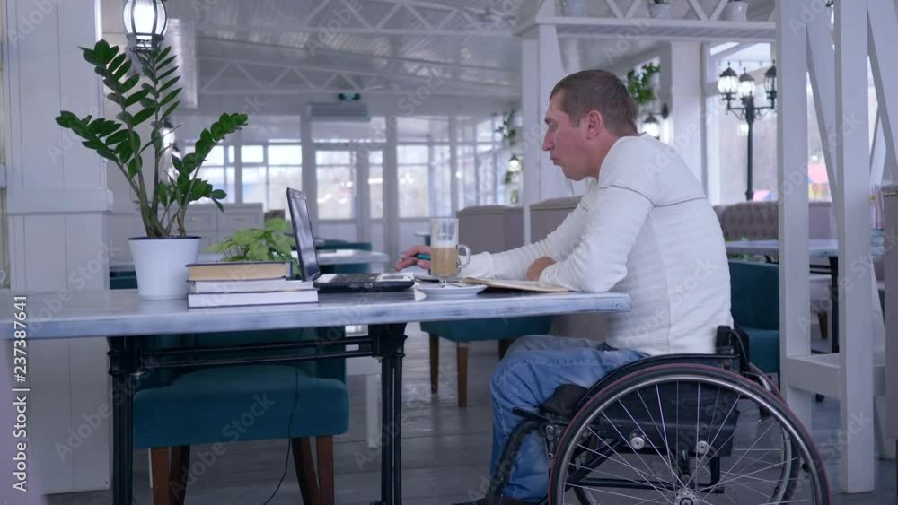 online courses, student crippled man in wheelchair uses a laptop and drinking coffee sitting at a table with books in a cafe online courses, student crippled man in wheelchair uses a laptop and drinking coffee sitting at a table with books in a cafe