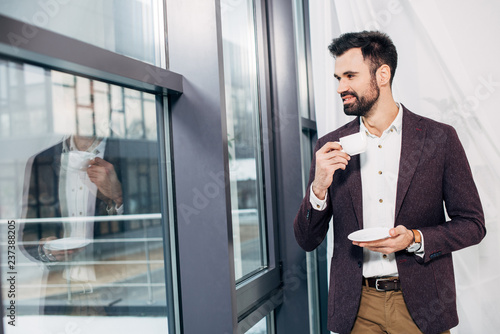 businessman looking at window, smiling and holding coffee cup in office