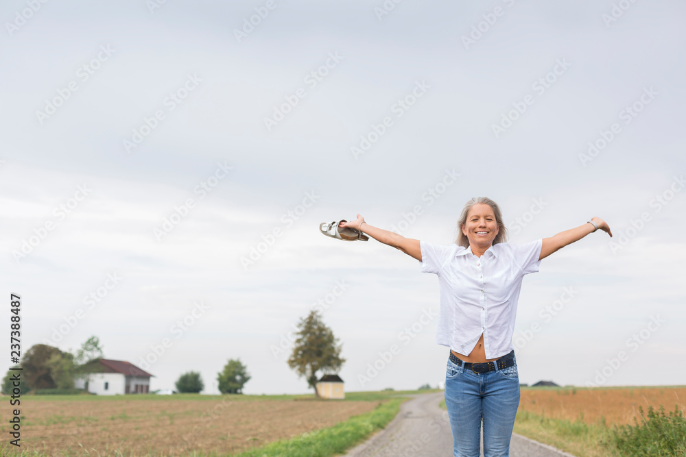 Mature woman standing on remote country lane in summer with outstretched arms Mature woman standing on remote country lane in summer with outstretched arms