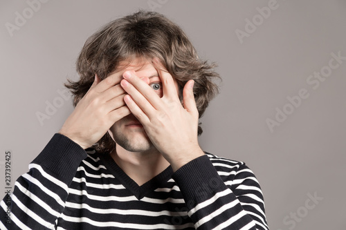Young hipster man peeking through fingers. Shaggy male covers face with both hands, has frightened expression as notices something terrible or scarying over grey background. Embarrassed expression