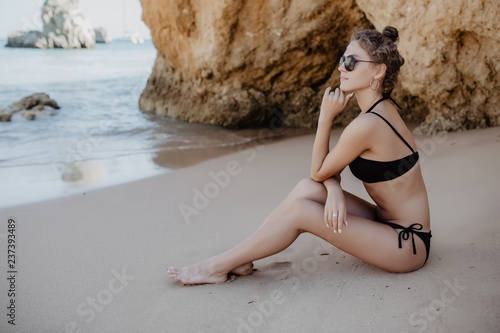 Portrait of young beautiful woman in bikini sitting at rocks near blue green clear ocean sea water. Sunbathing woman.