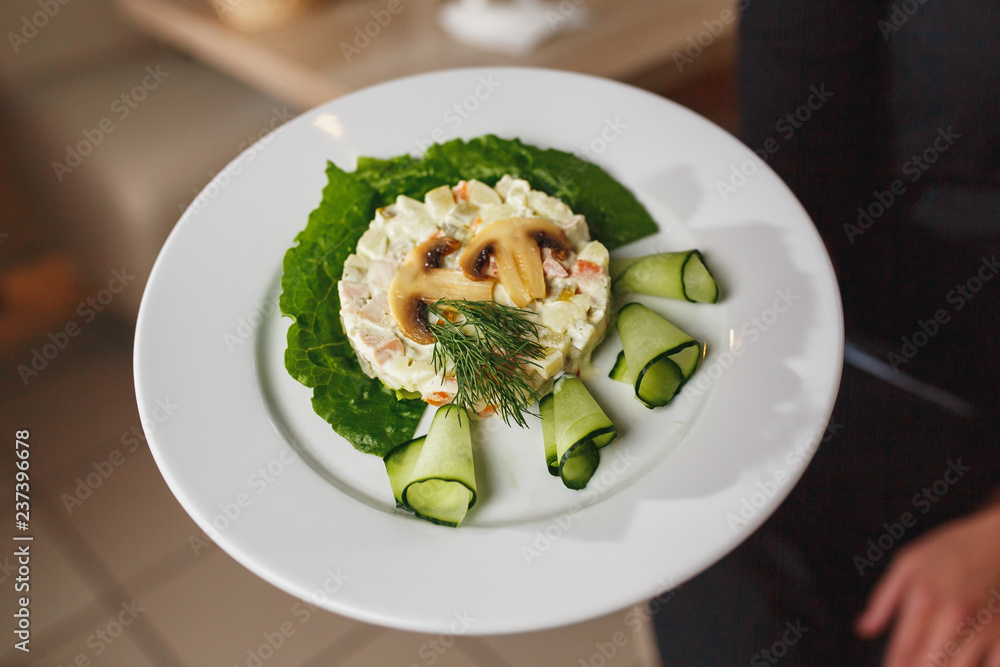 The waiter keeps the dish with Russian salad decorated with mushrooms and fresh cucumbers. Chef holding plate with tasty and delicious food and salad close up. The waiter keeps the dish with Russian salad decorated with mushrooms and fresh cucumbers. Chef holding plate with tasty and delicious food and salad close up.