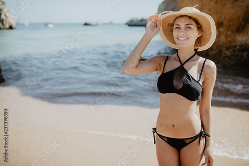 Happy smiling young woman in bikini and sunhat on sea beach