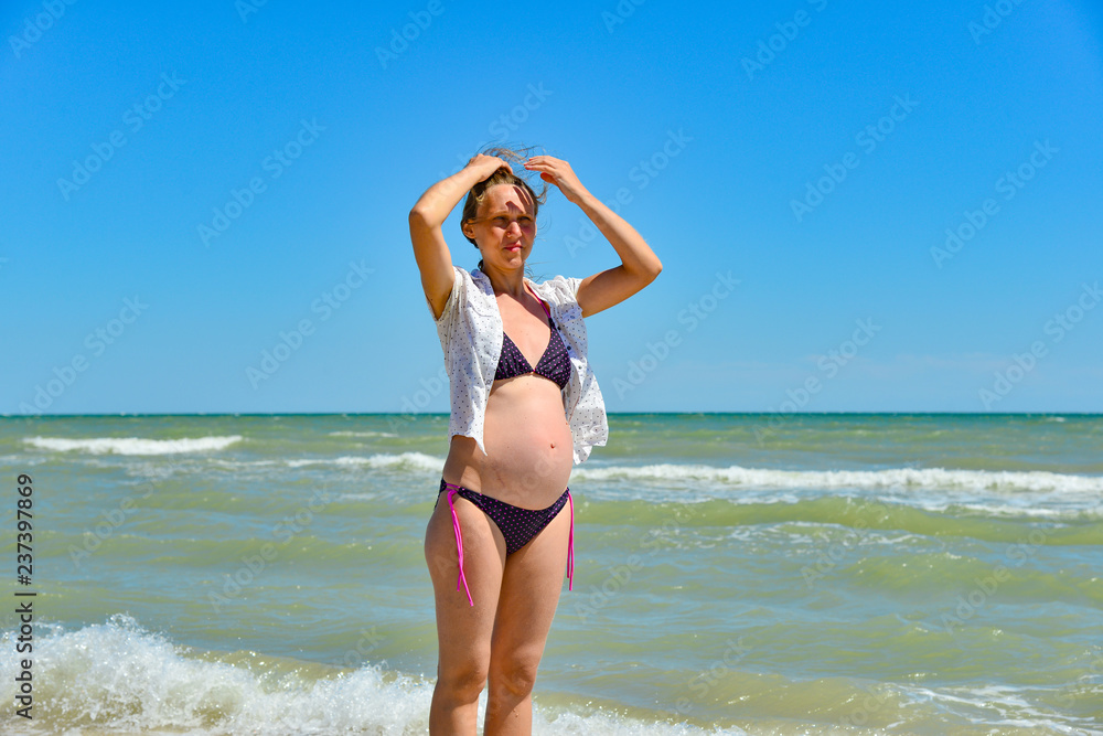 A happy and joyful pregnant woman stands by the sea and ocean on the beach under the sun in the open air, hair develops in the wind. Hands holding up and enjoying the sunshine.