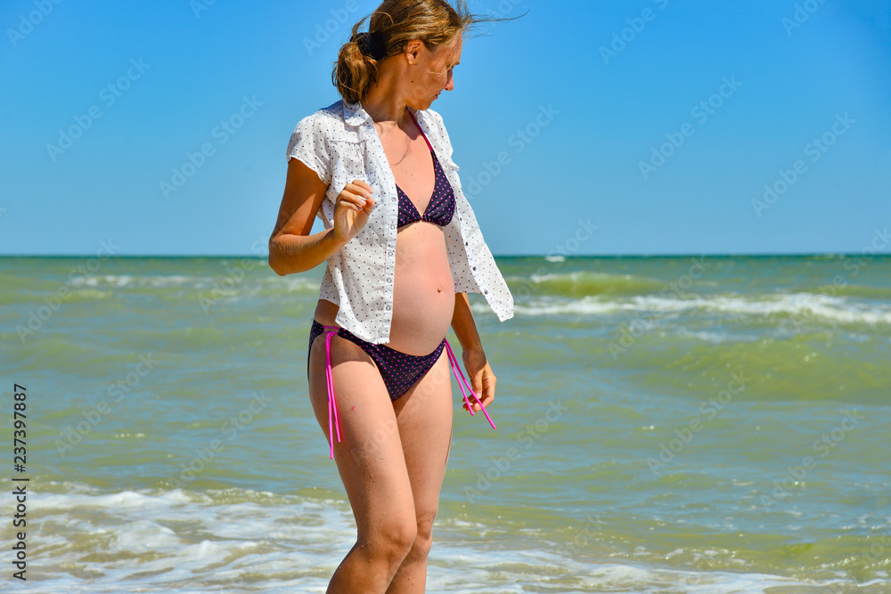 Naklejka premium A happy and joyful pregnant woman stands by the sea and ocean on the beach under the sun in the open air, hair develops in the wind.