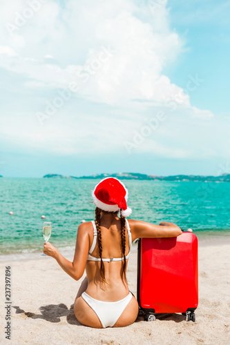 Beautiful Santa girl in bikini and Christmas hat on the beach of a tropical  island, in the hands of a glass of champagne and a red suitcase. Holiday  concept for the Christmas
