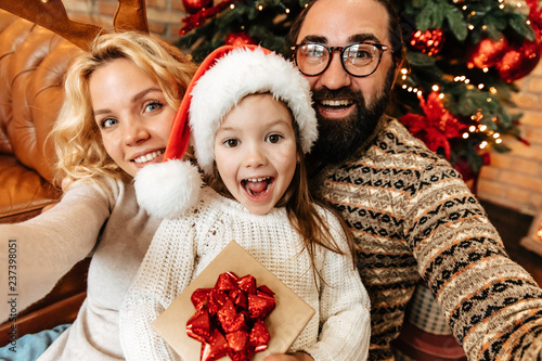 Christmas. Family. Home. Togetherness. Happy parents and their little girl are looking at camera and smiling near the Xmas tree photo